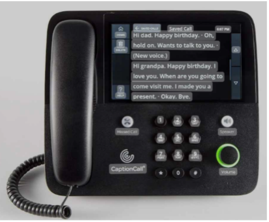 A CaptionCall phone displays a conversation transcript on its screen, showing a birthday message and other text. The phone has a handset, keypad, and buttons for call control, with a cord attached.