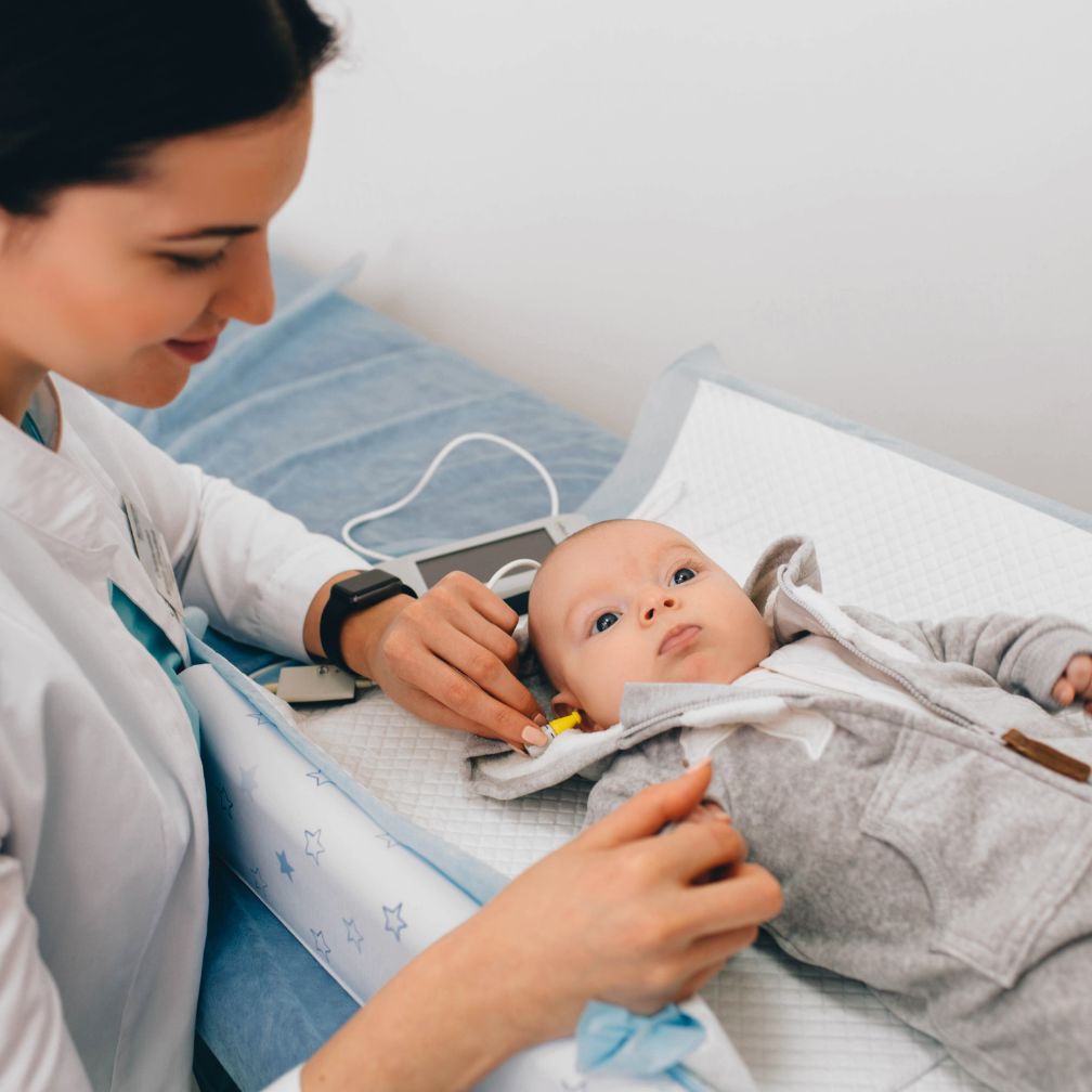A healthcare professional tests a babys hearing by placing small earphones in the babys ears while the baby lies on an exam table, looking up calmly.