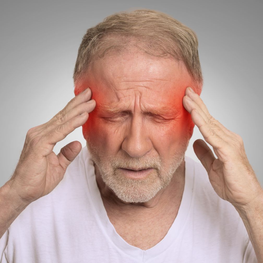 Older man with closed eyes holds his head with both hands, appearing to have a headache, with red highlighting around his temples to indicate pain or discomfort.