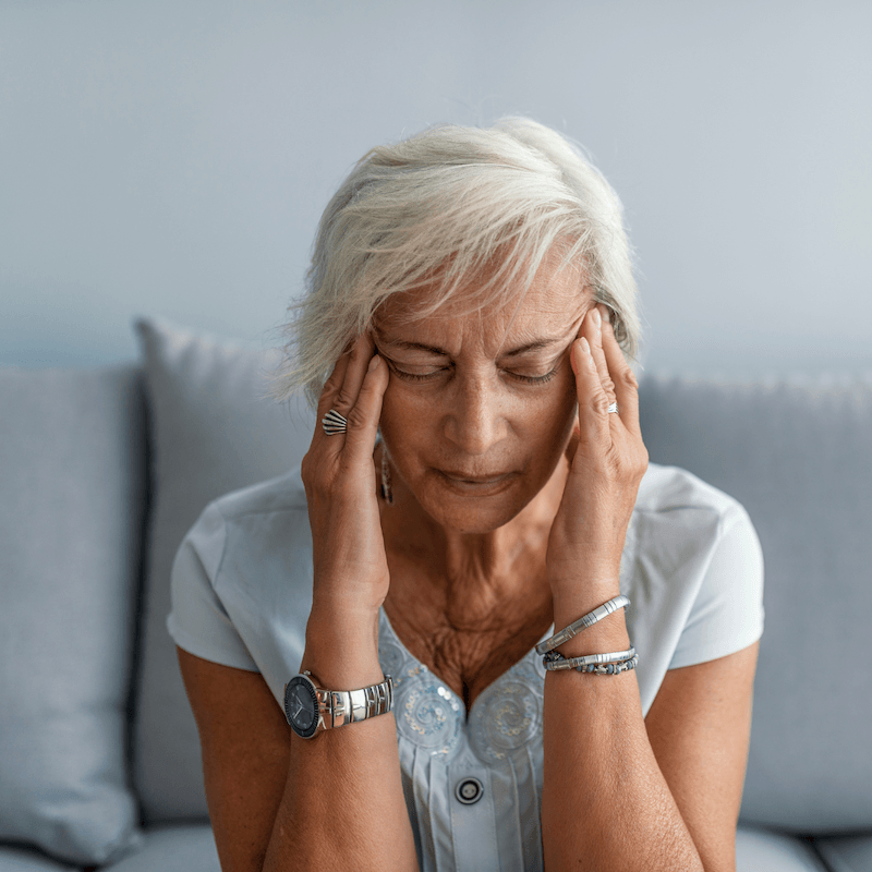 An older woman with short gray hair sits on a couch, eyes closed, and touches her temples with both hands, appearing to have a headache or feel stressed. She wears a light blue shirt and silver jewelry.