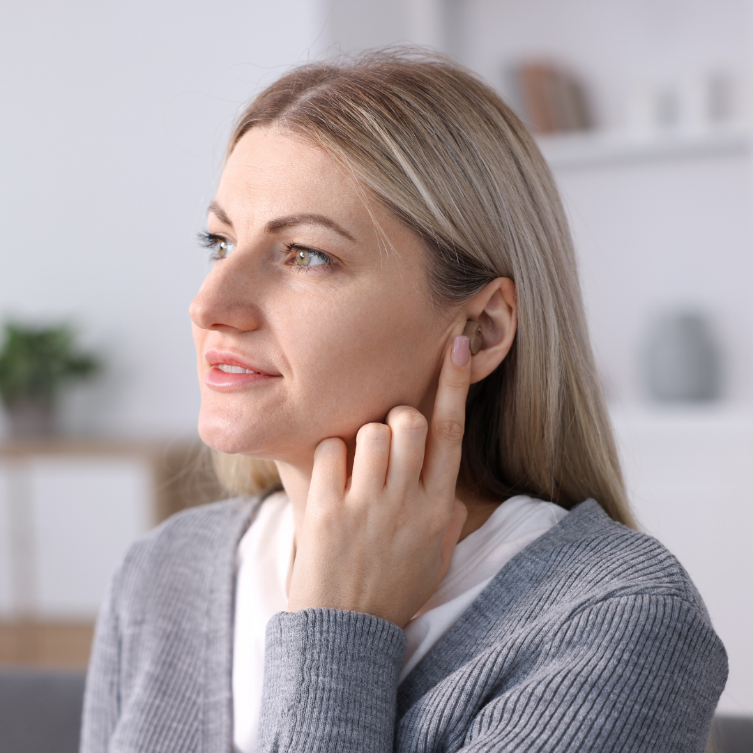 A woman with blonde hair touches her ear and looks to the left, appearing to listen attentively. She is wearing a gray sweater and is indoors in a softly lit room.