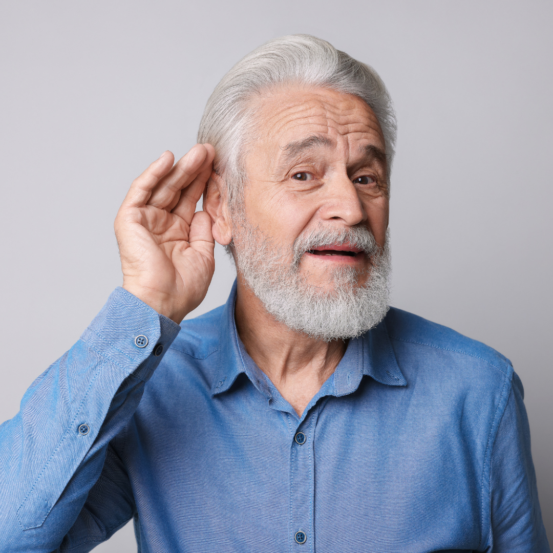 Older man with white hair and beard wearing a blue shirt, holding his hand to his ear as if trying to hear better, against a plain light background.
