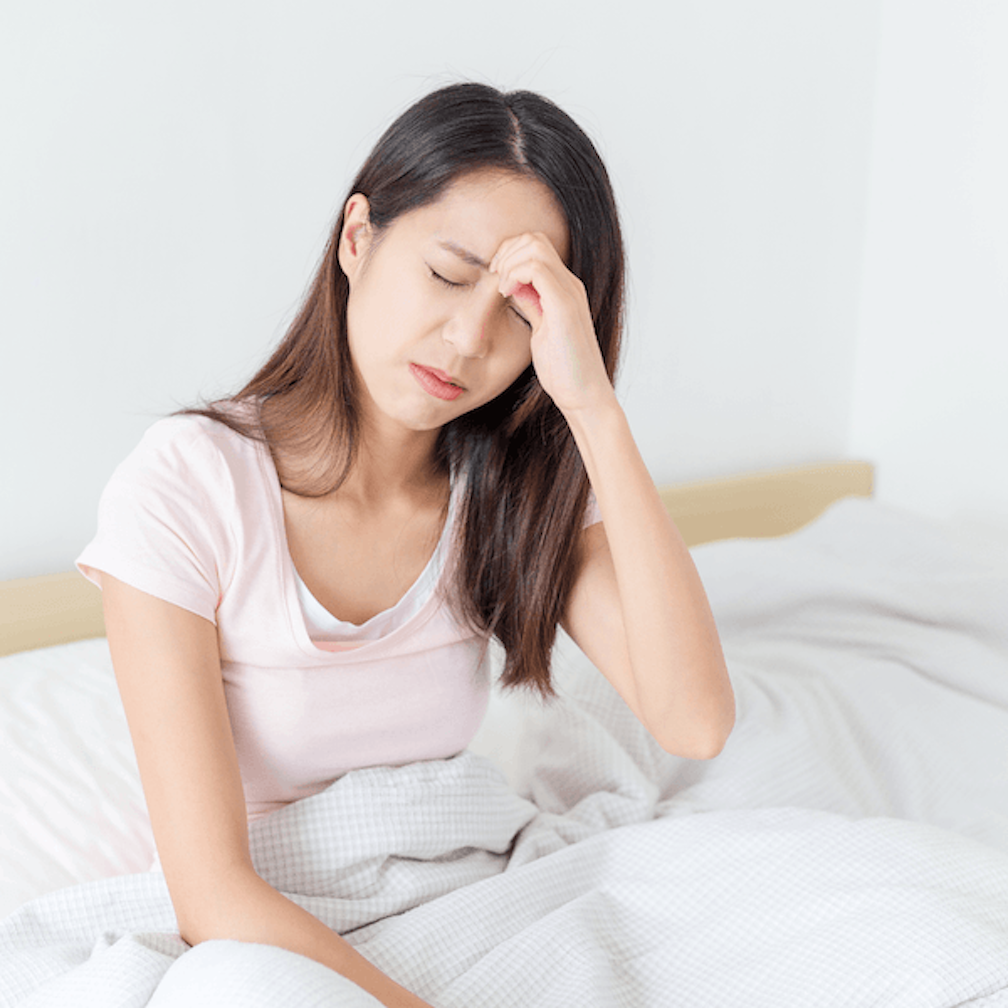 A young woman sitting in bed with her eyes closed, holding her forehead in pain.