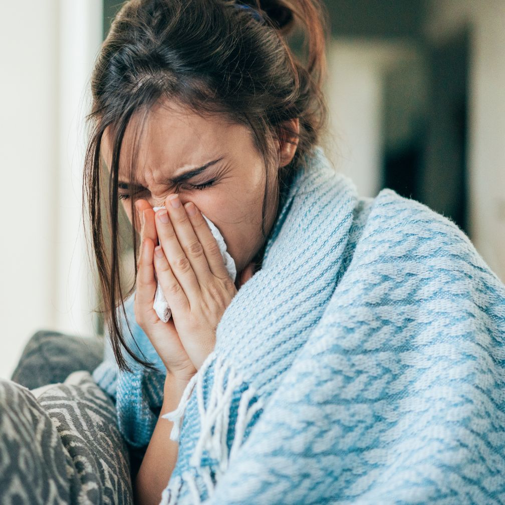 A young woman wrapped in a blue blanket sneezes into a tissue.