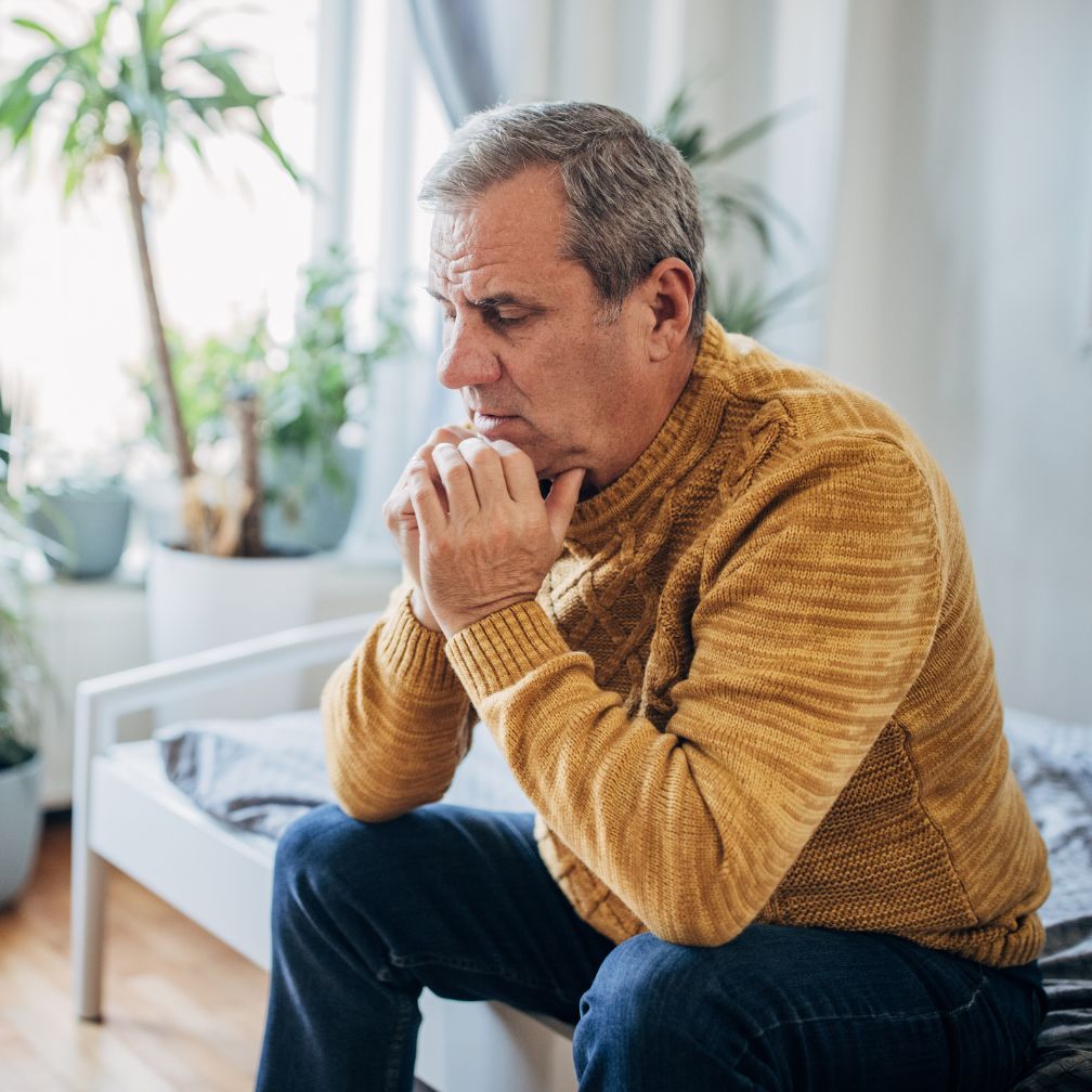 Middle-aged man sitting on the edge of a bed with his hands clasped under his chin, appearing deep in thought.